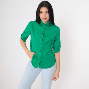 May include: A bright green button-down shirt with three-quarter sleeves. The shirt has a classic collar and is buttoned up. The model is wearing light blue jeans. The background is plain white.