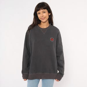 May include: Charcoal gray sweatshirt with a crew neck and a small red embroidered design on the chest. The sweatshirt has a darker gray trim at the neckline, cuffs, and hem. The model is wearing blue jeans.