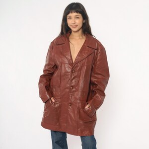 May include: A vintage brown leather jacket with a notched collar, three buttons, and two front pockets. The jacket is worn over blue jeans. The jacket appears to be a medium size.