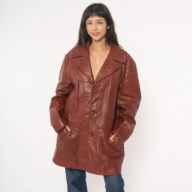 May include: A vintage brown leather jacket with a notched collar, three buttons, and two front pockets. The jacket is worn over blue jeans. The jacket appears to be a medium size.