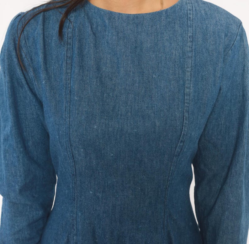 Puede incluir: Un vestido de mezclilla azul de manga larga con cuello redondo y detalles de costuras verticales que van desde los hombros hasta el corpi&ntilde;o, creando un ajuste estructurado en el torso con una textura de tela de lavado medio.
