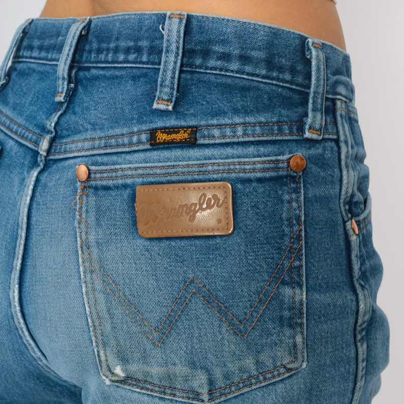 May include: Close-up of blue denim jeans with a Wrangler logo on the back pocket. The jeans have copper rivets and a small Wrangler label. The denim has a faded, worn appearance, typical of vintage jeans.