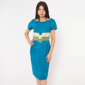 Könnte beinhalten: Ein knielanges blaues Kleid mit kurzen Ärmeln und einem Rundhalsausschnitt. Der Taillenbereich ist mit einem horizontalen beigen Einsatz und einem grünen Einsatz akzentuiert, die jeweils mit einem passenden runden Knopf verziert sind.