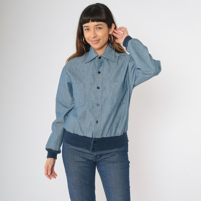 May include: A blue denim shirt jacket with a button-up front and a navy blue ribbed knit waistband.
