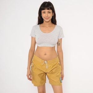 May include: A woman wearing a gray crop top and mustard yellow shorts with a drawstring waist and side pockets. The shorts have a small patch on the lower right leg. The woman is smiling at the camera.