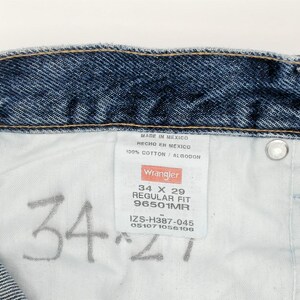 May include: Close-up of the inside of blue denim jeans. The jeans have a white tag with the Wrangler logo and size information: 34 x 29 Regular Fit. The jeans are made of 100% cotton.