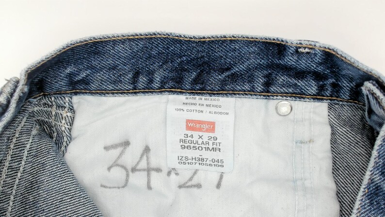 May include: Close-up of the inside of blue denim jeans. The jeans have a white tag with the Wrangler logo and size information: 34 x 29 Regular Fit. The jeans are made of 100% cotton.