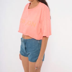 K&ouml;nnte beinhalten: Ein neonfarbenes, bauchfreies T-Shirt mit kurzen &Auml;rmeln und Rundhalsausschnitt, das auf der Brust den Schriftzug BON JON in einem helleren Farbton zeigt, kombiniert mit einer hoch taillierten blauen Jeansshorts mit Knopf- und Rei&szlig;verschluss.