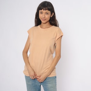 May include: A peach-colored, short-sleeved t-shirt. The shirt has a crew neck and a relaxed fit. The model is wearing blue jeans. The background is plain white. The shirt is made of a soft, comfortable material.