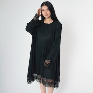 Puede incluir: Un vestido negro de manga larga con un borde de encaje alrededor del dobladillo y los puños. El vestido tiene un frente abotonado y un ajuste holgado.