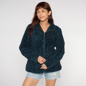 Puede incluir: Chaqueta de forro polar de color turquesa con cierre de cremallera completo. La chaqueta presenta un estampado de cuadros en el cuello y un pequeño bolsillo. La chaqueta es de un material suave y texturizado.