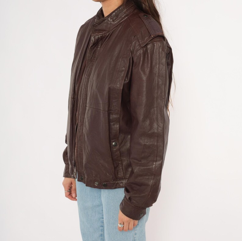 May include: A brown leather jacket with a stand-up collar and button details. The jacket has a zip closure and shoulder epaulettes. The sleeves are long, and the jacket appears to be vintage.