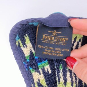 May include: A dark blue, green, and cream patterned cotton towel with a dark blue fabric border. A black rectangular tag is attached, featuring the text PENDLETON HOME COLLECTION, 100% COTTON, 100% COTON, MADE IN CHINA, and FABRIQUE EN CHINE.