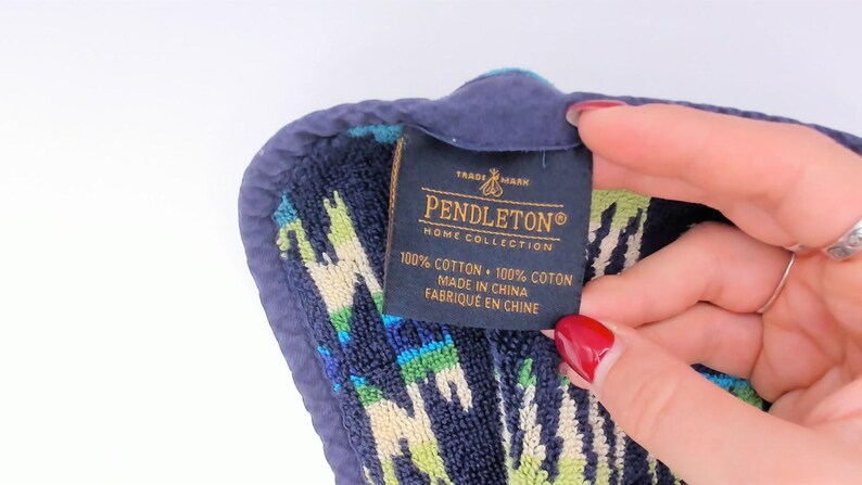 May include: A dark blue, green, and cream patterned cotton towel with a dark blue fabric border. A black rectangular tag is attached, featuring the text PENDLETON HOME COLLECTION, 100% COTTON, 100% COTON, MADE IN CHINA, and FABRIQUE EN CHINE.