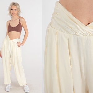 May include: Cream-colored, high-waisted pants with a unique, twisted waistband. The pants have a loose, flowing fit and are paired with a brown crop top. The model is wearing white sneakers. The pants are made of a soft, lightweight fabric.