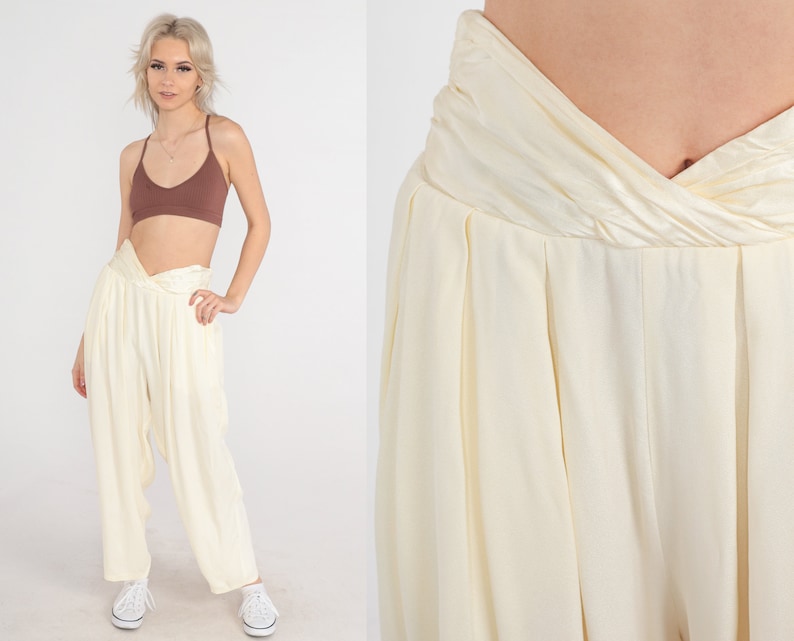 May include: Cream-colored, high-waisted pants with a unique, twisted waistband. The pants have a loose, flowing fit and are paired with a brown crop top. The model is wearing white sneakers. The pants are made of a soft, lightweight fabric.