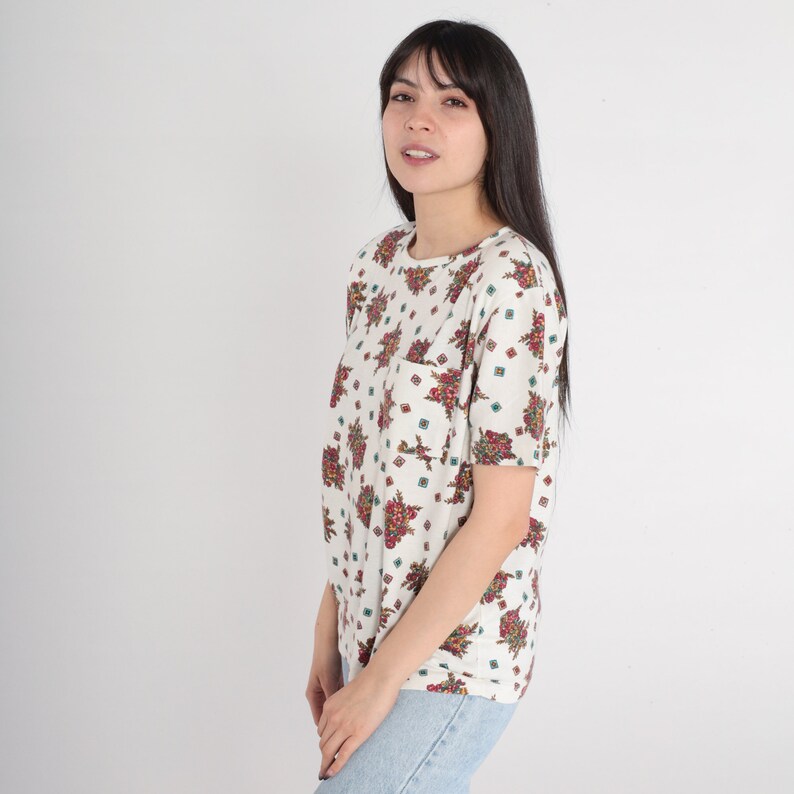 May include: White short-sleeved shirt with a floral pattern in shades of red, green, and blue. The shirt has a pocket on the left side.