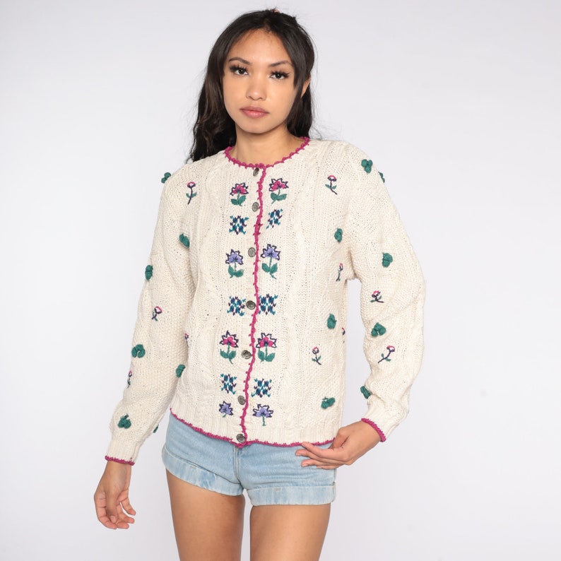 May include: A cream-colored knitted cardigan with a floral embroidered design. The cardigan has a pink button closure and a white crocheted trim.