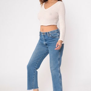 May include: A woman wearing a white cropped sweater and blue denim jeans with frayed hems. She is wearing white sneakers.
