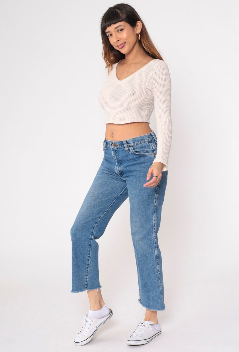 May include: A woman wearing a white cropped sweater and blue denim jeans with frayed hems. She is wearing white sneakers.