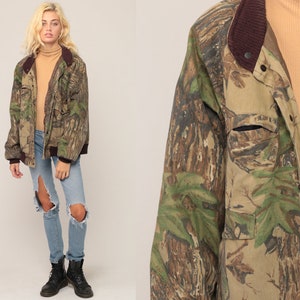 Op de afbeelding: Een jas met camouflageprint, voorzien van een donkerbruine geribbelde kraag en boorden, een ritssluiting aan de voorzijde en een borstzak. De jas wordt gedragen over een beige coltrui, een lichtblauwe gescheurde spijkerbroek en zwarte veterschoenen.