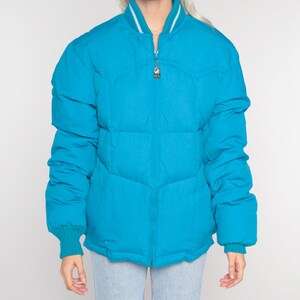May include: A turquoise blue puffer jacket with a white ribbed collar and cuffs. The jacket has a zipper closure and a quilted design.
