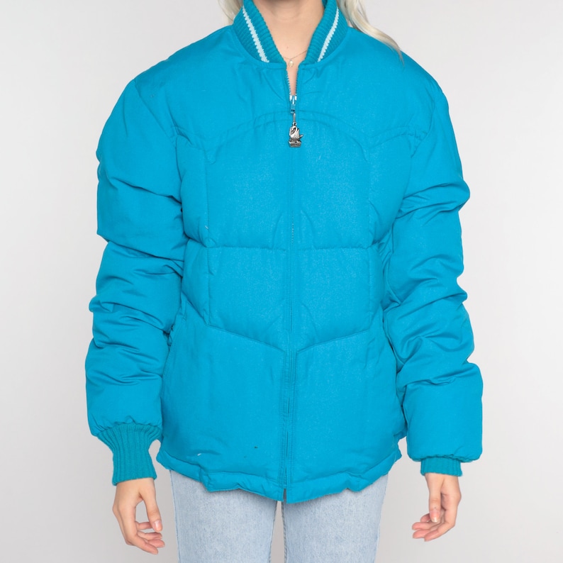 May include: A turquoise blue puffer jacket with a white ribbed collar and cuffs. The jacket has a zipper closure and a quilted design.