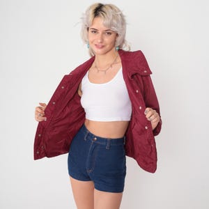May include: A burgundy quilted jacket with a high collar and snap closures. The jacket is open, revealing a white crop top and high-waisted denim shorts. The outfit is styled with a silver necklace and turquoise earrings.