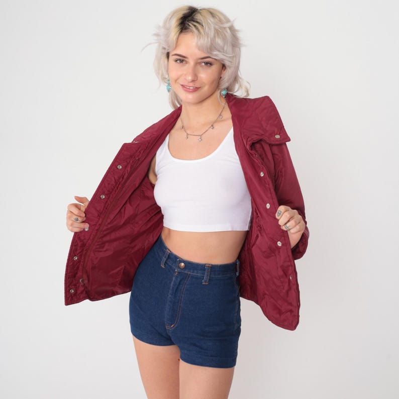 May include: A burgundy quilted jacket with a high collar and snap closures. The jacket is open, revealing a white crop top and high-waisted denim shorts. The outfit is styled with a silver necklace and turquoise earrings.