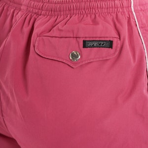 May include: Close-up of pink shorts with a white stripe along the side. The shorts have an elasticated waistband and a back pocket with a silver button and a black label that reads "AMBCO".