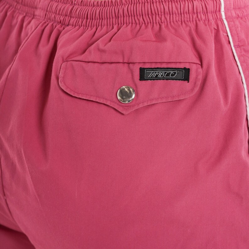 May include: Close-up of pink shorts with a white stripe along the side. The shorts have an elasticated waistband and a back pocket with a silver button and a black label that reads "AMBCO".
