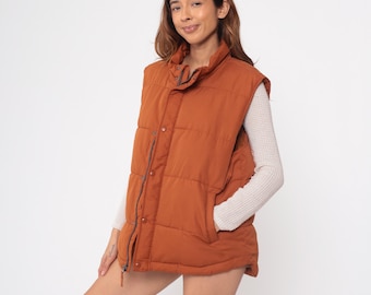 May include: A rust-colored puffer vest with a high collar and a zipper closure. The vest has a quilted design and two front pockets. The model is wearing a long-sleeved, off-white shirt underneath the vest.