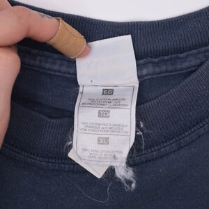 May include: A white tag with black text on a dark blue t-shirt. The tag shows the size XL and the fabric content: 100% ring spun cotton, made in Mexico. The tag also includes care instructions in Spanish and French.