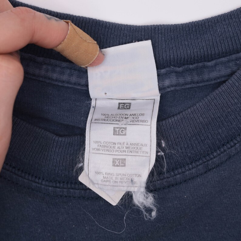 May include: A white tag with black text on a dark blue t-shirt. The tag shows the size XL and the fabric content: 100% ring spun cotton, made in Mexico. The tag also includes care instructions in Spanish and French.