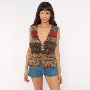 May include: A sleeveless knit vest featuring horizontal stripes in shades of red, black, and tan with a textured, multi-colored yarn finish. The garment has a deep V-neckline and is secured at the front with three light-colored wooden toggle buttons.