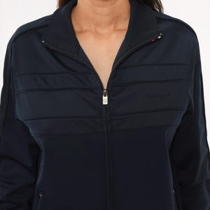 May include: A dark navy blue zip-up track jacket featuring a high collar, long sleeves, and horizontal quilted panel detailing across the chest. The jacket includes a central silver-toned zipper pull and subtle tonal branding on the right side.