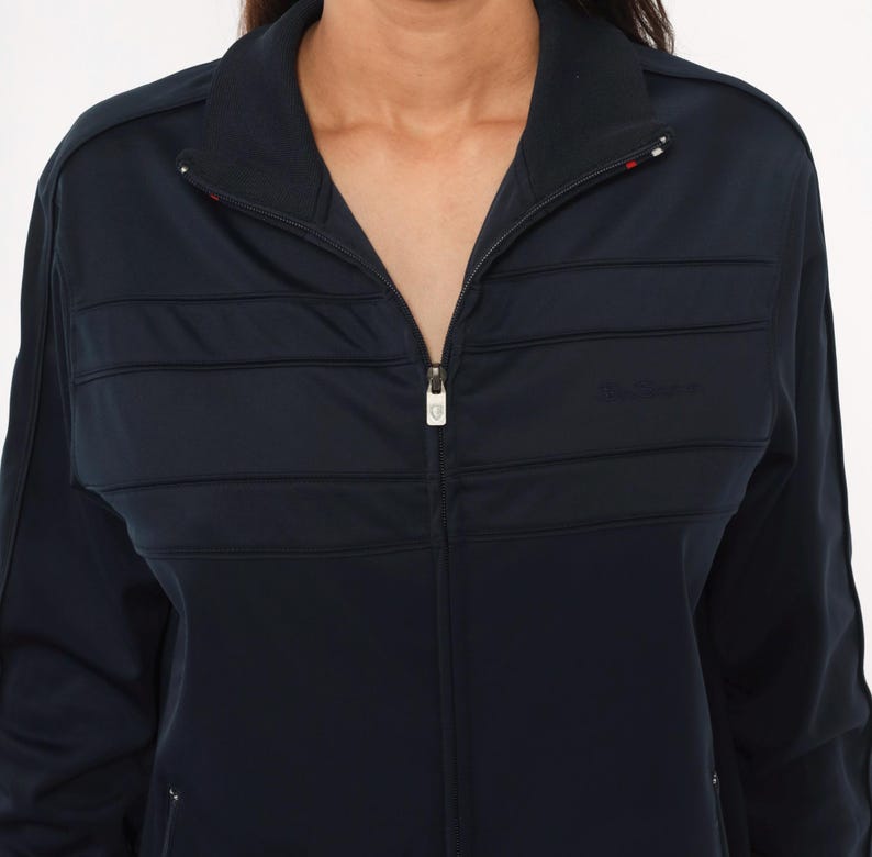 May include: A dark navy blue zip-up track jacket featuring a high collar, long sleeves, and horizontal quilted panel detailing across the chest. The jacket includes a central silver-toned zipper pull and subtle tonal branding on the right side.