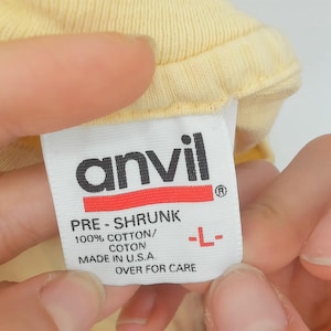 May include: A close-up of a yellow garment tag held by a person. The tag is white with the black "anvil" logo and red stripe. Text includes "PRE-SHRUNK", "100% COTTON/COTON", "MADE IN U.S.A", and size "L-".