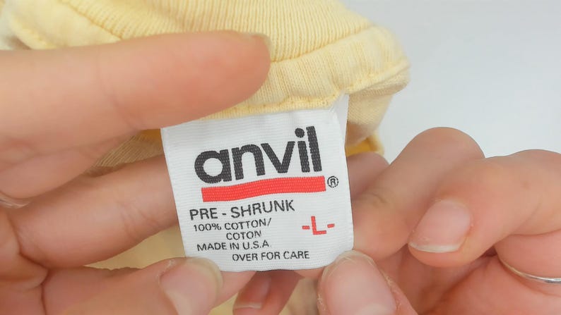 May include: A close-up of a yellow garment tag held by a person. The tag is white with the black "anvil" logo and red stripe. Text includes "PRE-SHRUNK", "100% COTTON/COTON", "MADE IN U.S.A", and size "L-".