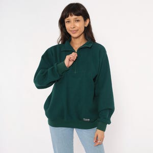 May include: A dark green fleece pullover with a quarter-zip neckline. The long-sleeved top has a relaxed fit, a small rectangular patch near the hem, and ribbed cuffs and hem. The model is wearing light blue jeans.