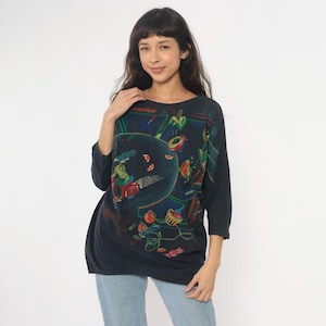 May include: Dark navy blue long-sleeve shirt with a colorful abstract design. The artwork features a circular shape with figures and fruit-like objects in shades of red, green, yellow, and blue. The shirt has a round neckline and three-quarter sleeves.