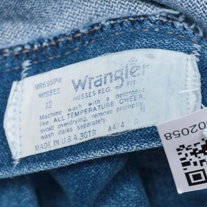 Op de afbeelding: Een blauw denim label met witte tekst die "Wrangler Misses Reg. Fit" en "Machine wash with a detergent like ALL TEMPERATURE CHEER, avoid overdrying, remove promptly, wash darks separately. Made in U.S.A. 3GTR A4/4 R" luidt.