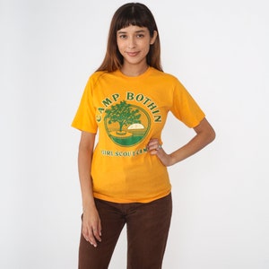 May include: Mustard yellow t-shirt with a green and white graphic that reads "CAMP BOTHIN GIRL SCOUT CAMP". The graphic features a tree, sun, and water design. The shirt is a short-sleeved, crew neck style.