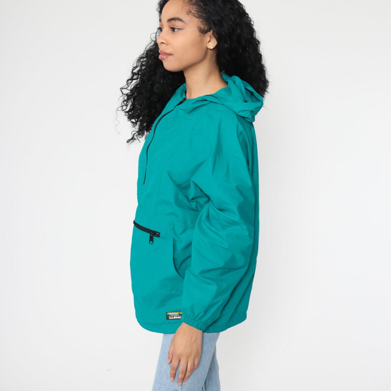 May include: Teal blue hooded jacket with a zipper closure and a large front pocket. The jacket has a drawstring hood and a logo patch on the bottom left side.