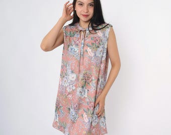 May include: Sleeveless, knee-length dress in a soft peach hue, adorned with a floral pattern in shades of blue, white, and green. The dress features a tie-neck detail and a relaxed, flowing silhouette, perfect for warm weather.