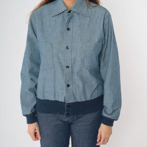 May include: A blue denim shirt with a button-up front and a navy blue ribbed knit band at the bottom.