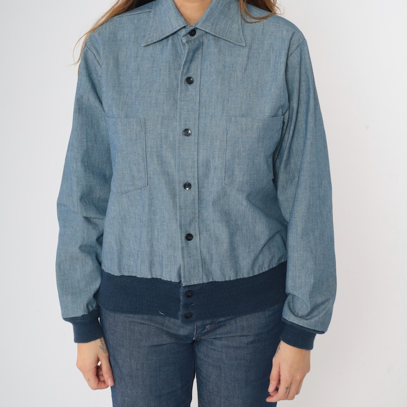 May include: A blue denim shirt with a button-up front and a navy blue ribbed knit band at the bottom.
