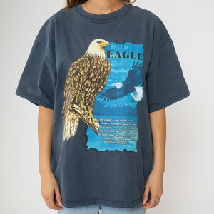 May include: A dark blue t-shirt featuring a graphic print of a bald eagle perched on a branch. The text "BALD EAGLE" is printed above the eagle. The text below the eagle reads: "The Eagle is one of the world's largest and most majestic birds of prey. One of only two kinds of eagles breed in North America, the bald eagle is grounded by feathers for the grandeur of the Pacific Northwest."