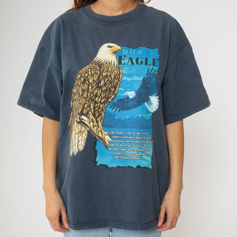May include: A dark blue t-shirt featuring a graphic print of a bald eagle perched on a branch. The text "BALD EAGLE" is printed above the eagle. The text below the eagle reads: "The Eagle is one of the world's largest and most majestic birds of prey. One of only two kinds of eagles breed in North America, the bald eagle is grounded by feathers for the grandeur of the Pacific Northwest."