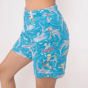 K&ouml;nnte beinhalten: Blaue Badeshorts mit einem Muster aus Cartoon-Surfern und Haien. Die Shorts haben einen Kordelzug.